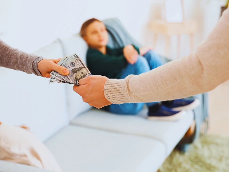 Handing over cash with a child in the background. Parenting Time Reduces Child Support