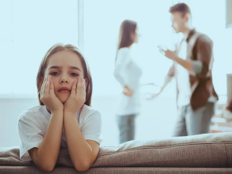 A sad little blond girl dressing a white shirt with his parents fighting behind her Used Against You in an Illinois Child Custody Case
