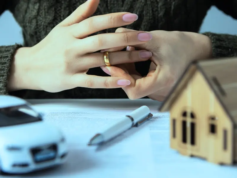 A close-up of a woman's hands removing her wedding ring. In front of her are a house and a car, indicating a separation of assets is imminent. private equity investments in an Illinois divorce