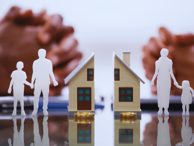 A miniature house split in half, alongside miniature people. In the background, two people with their hands crossed. standard of living after an Illinois divorce, budgeting after divorce